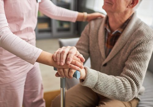 Close-up of young caregiver holding hands with elderly man and talking to him, she caring about him at home